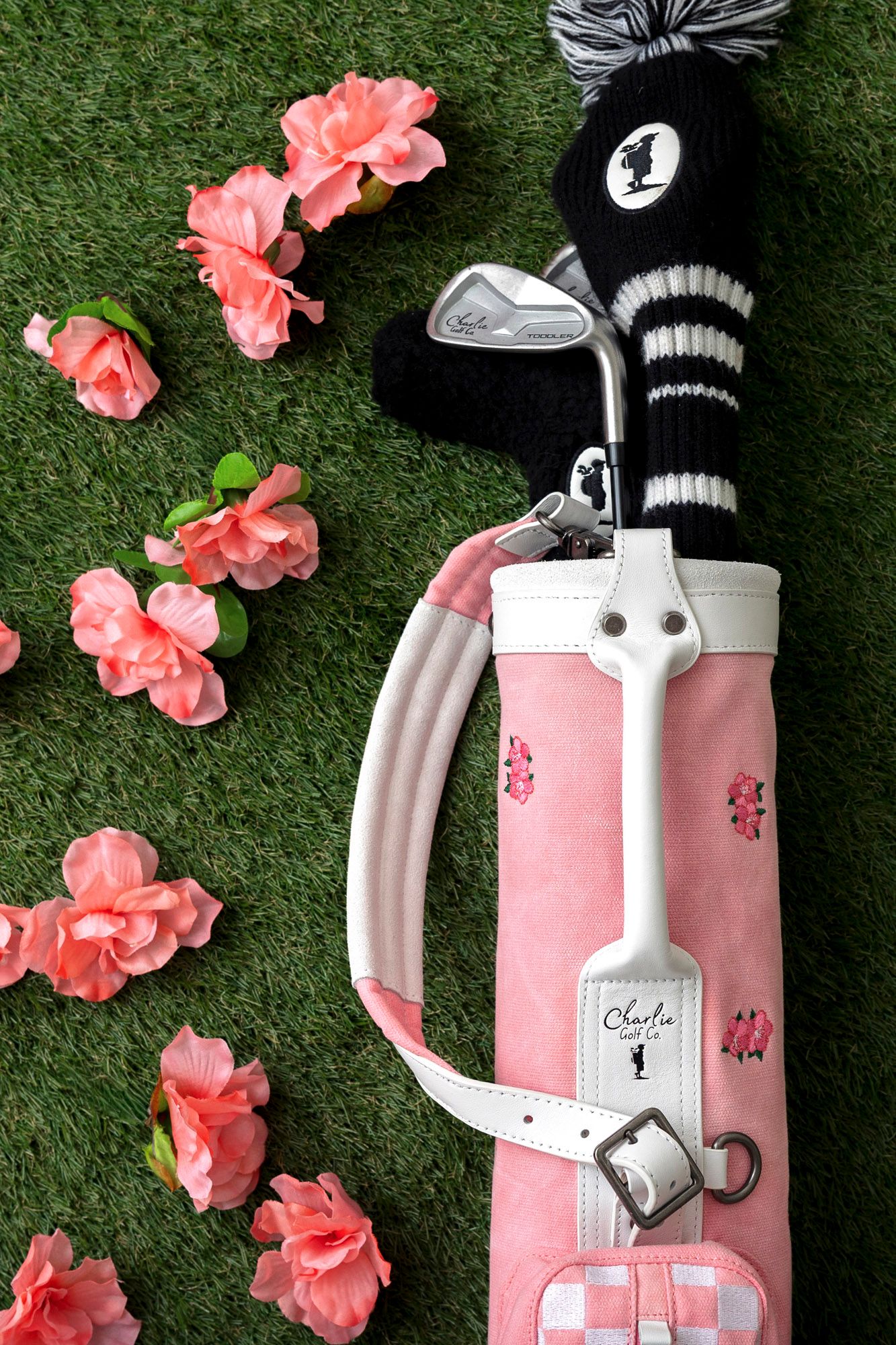 Pink vintage golf bag with black pom pom headcover displayed on grass with scattered coral flowers.
