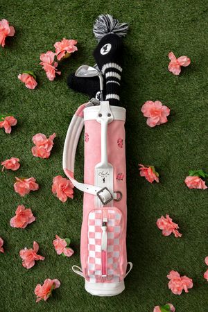 Pink golf bag with checkered pattern lies on green grass surrounded by scattered coral-colored flowers.