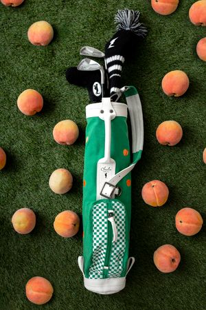 A green golf bag with checkered pattern lies on grass surrounded by scattered orange peaches.