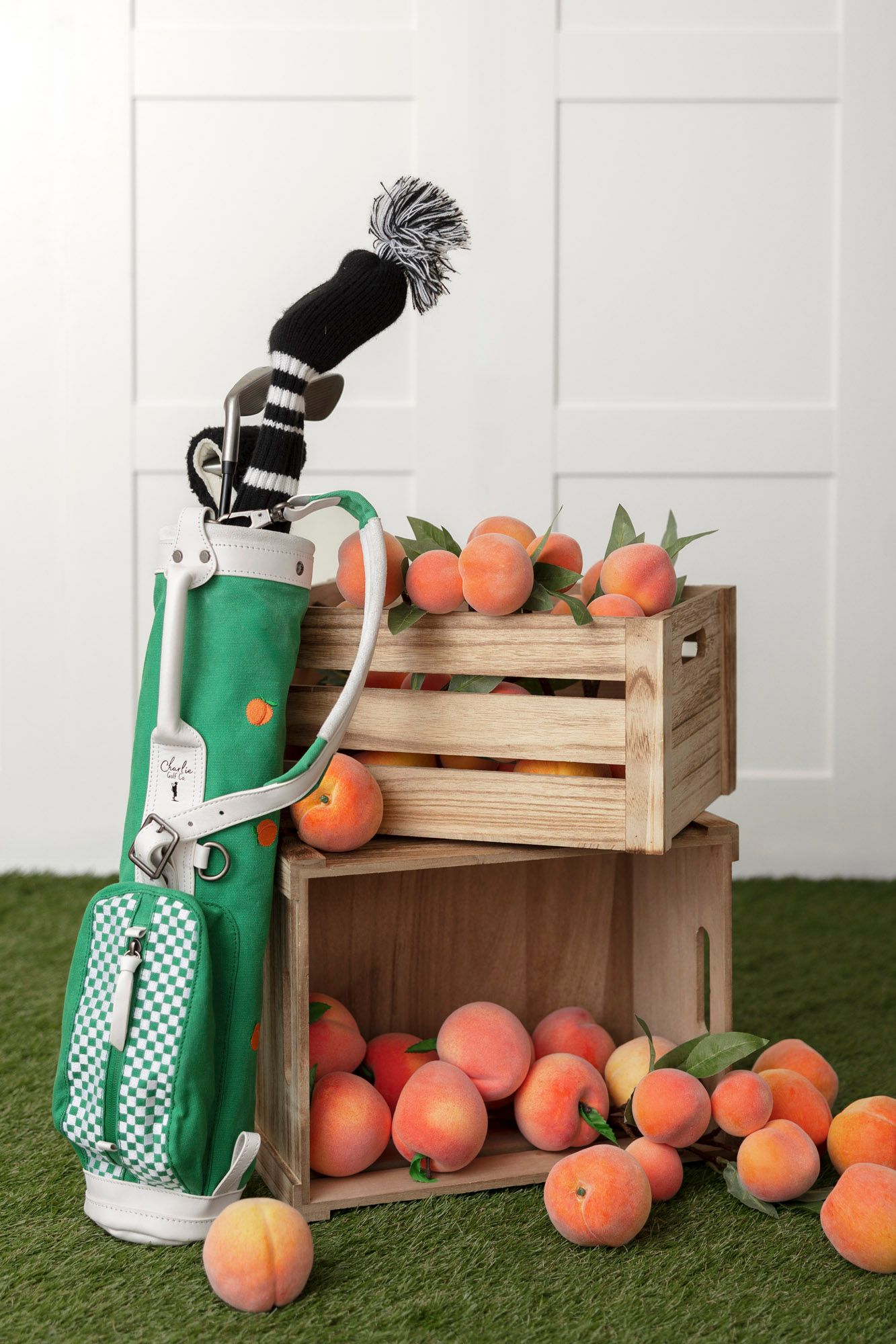 Retro green golf bag next to wooden crate filled with fresh peaches on artificial turf.
