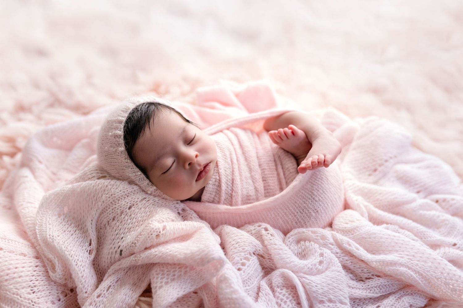 A sleeping newborn baby wrapped in soft pink blankets and lace on a delicate pink textured background.