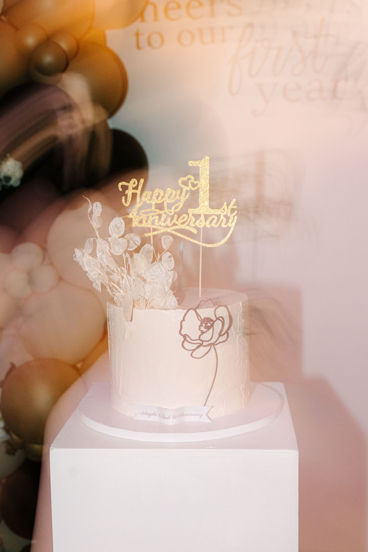 White wedding cake with delicate gold anniversary cake topper decorations against a soft blush pink background.