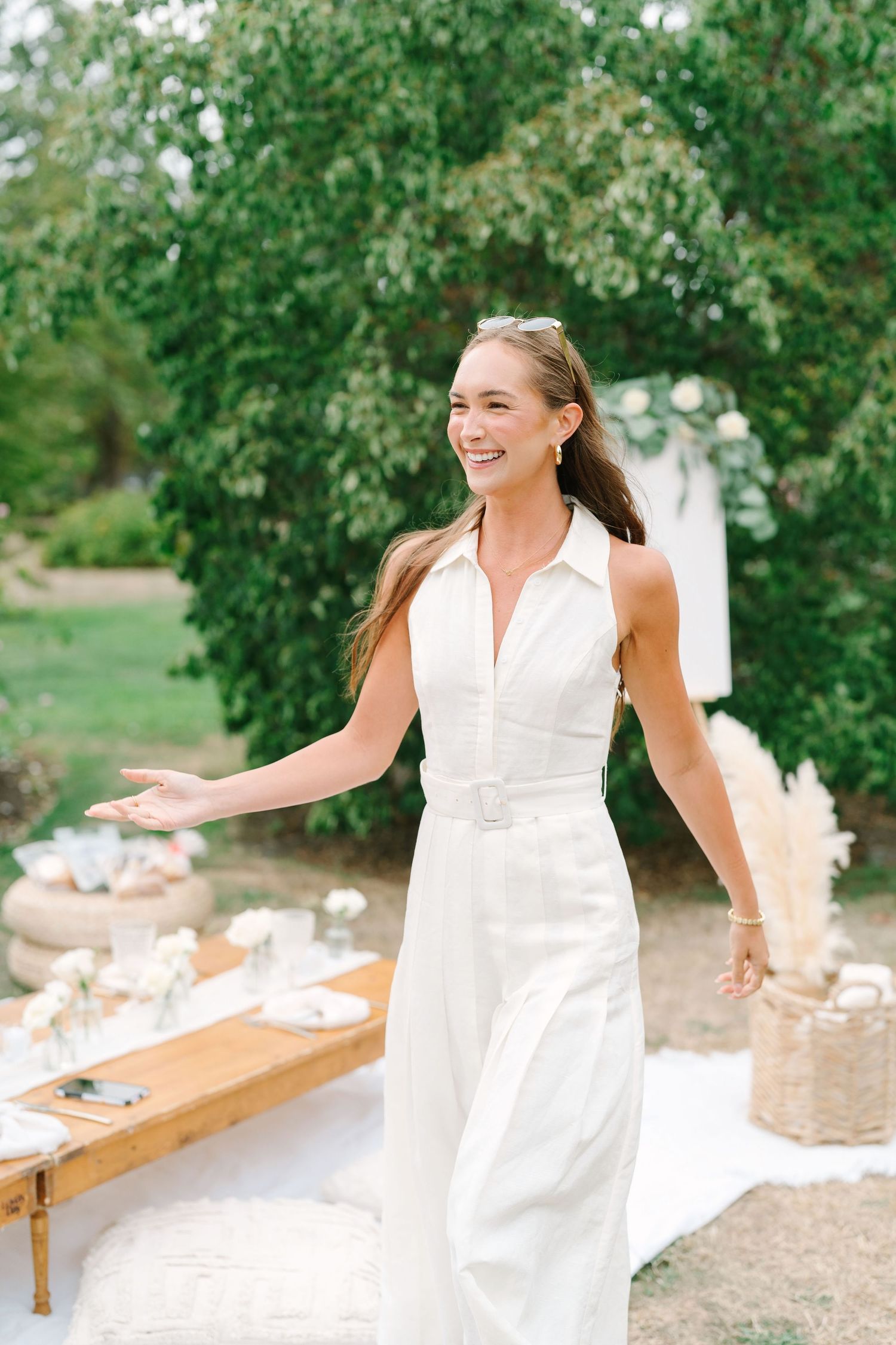A person in a flowing white dress laughs joyfully while sitting on a wooden bench in a garden setting.