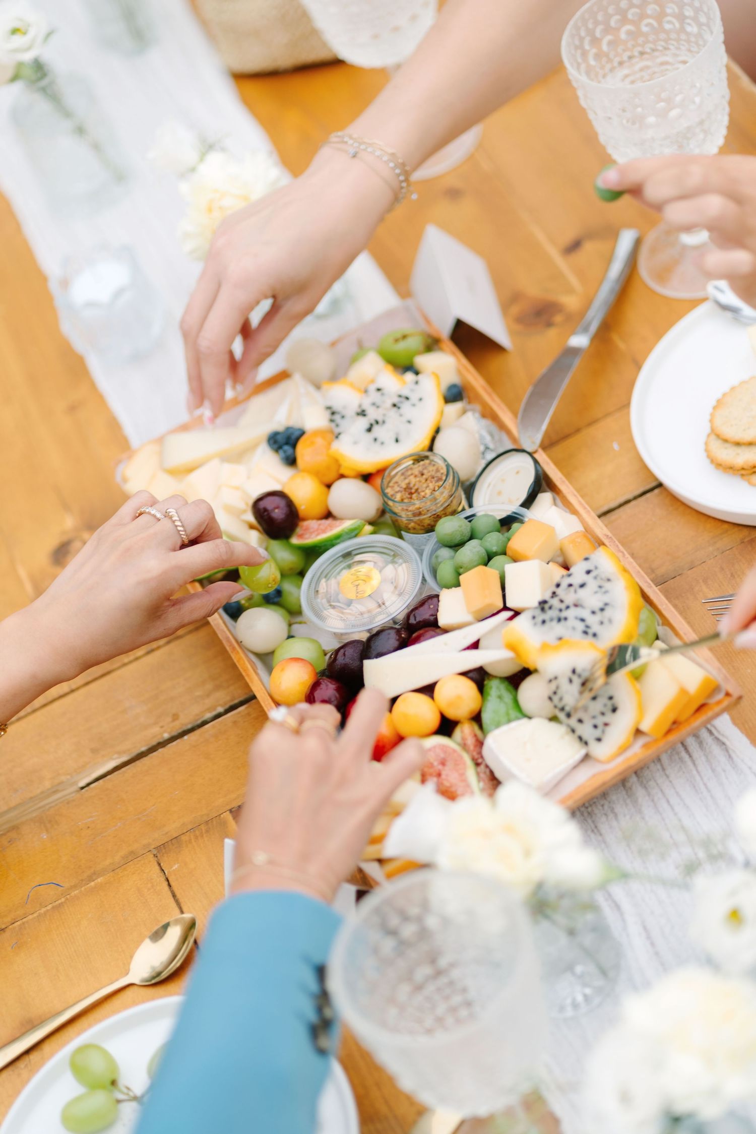 Hands reaching for appetizers and snacks on a wooden table with various cheeses and fresh vegetables.