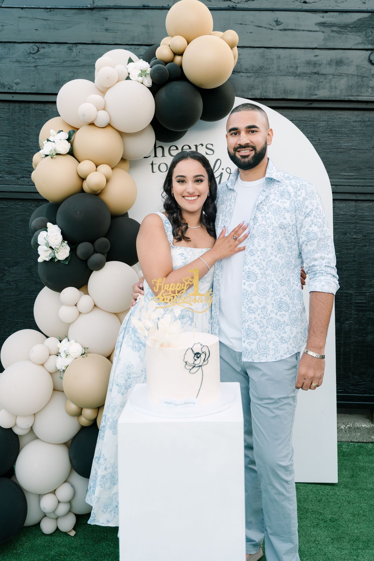 A couple in coordinating white outfits poses in front of a balloon arch and floral decorations at a celebration.