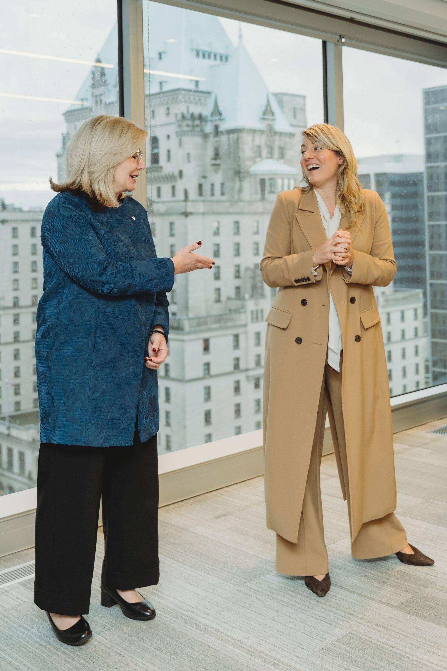 Two people in business attire share a laugh while standing on a balcony overlooking city buildings.