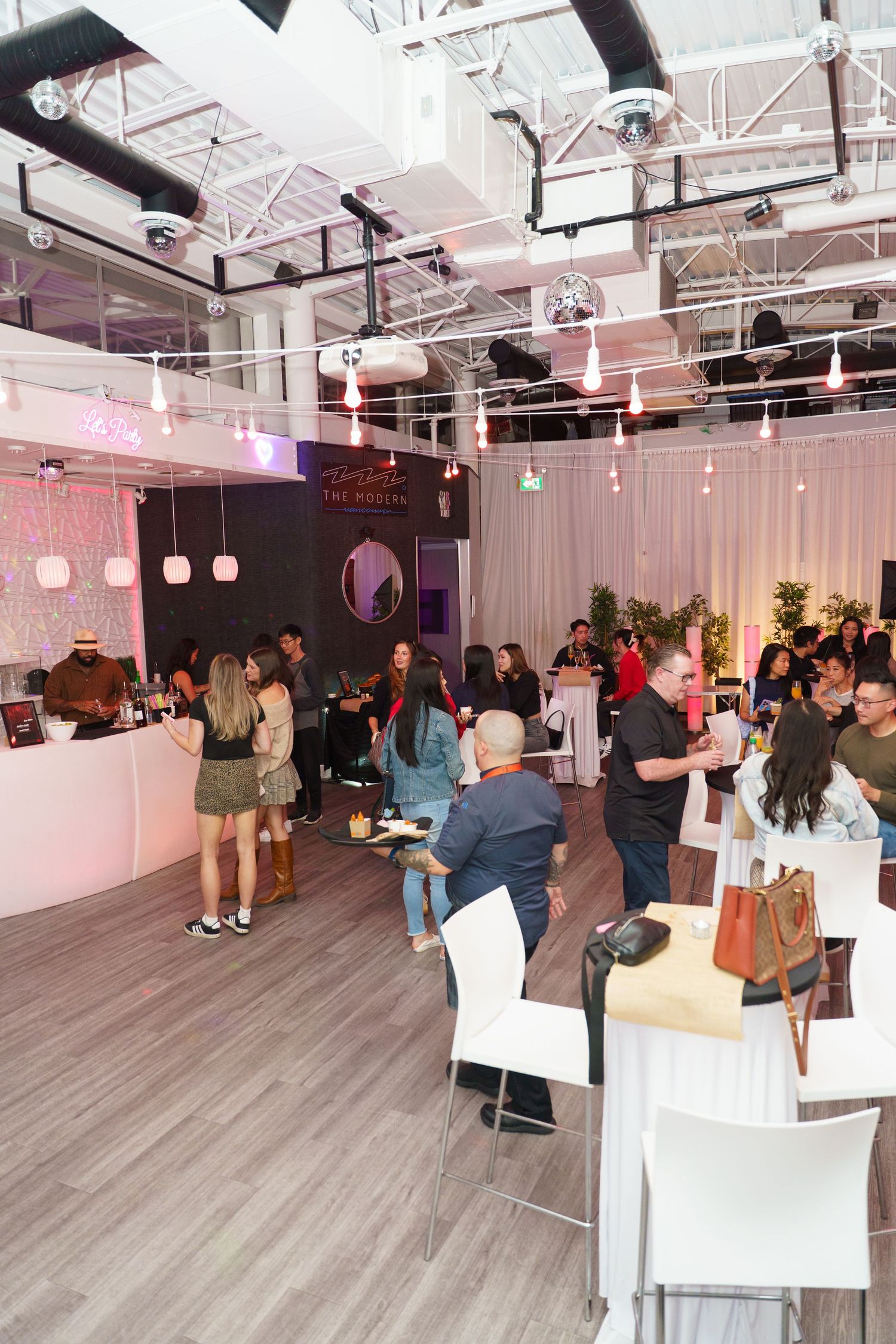 Modern event space with pink lighting features guests mingling in an open floor plan with white decor and industrial ceiling.