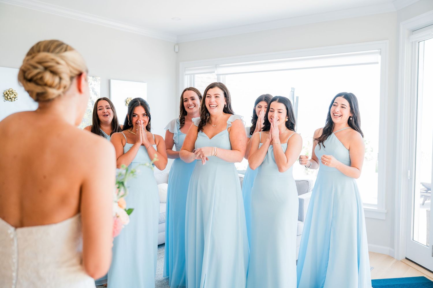Bridesmaids in flowing light blue dresses react with joy and excitement during a wedding preparation moment.