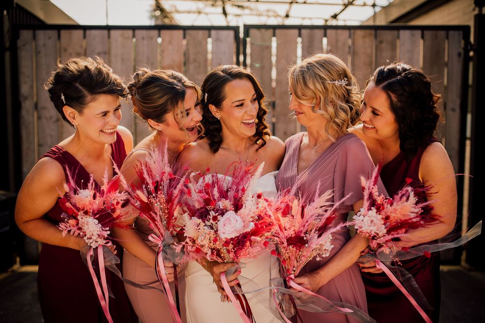 Bridesmaids in formal attire lean in together holding pink feather fans at a wedding celebration.
