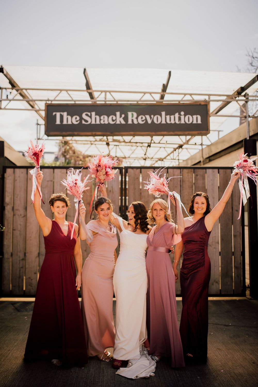 A wedding party in burgundy and pink dresses poses together at sunset against a rustic wooden backdrop.