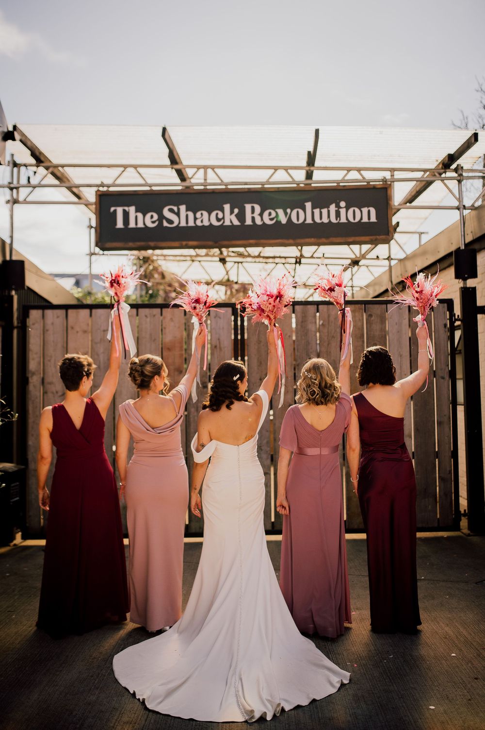 Wedding party in burgundy dresses and white gown pose with raised arms against rustic backdrop at sunset.
