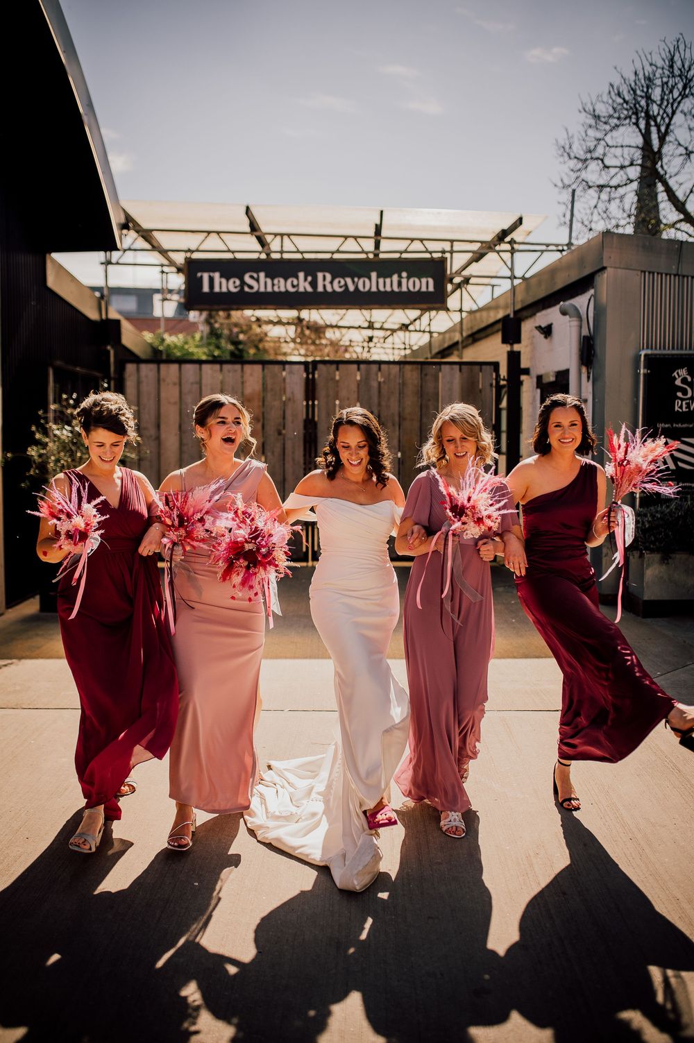 Wedding party in burgundy and pink dresses poses together casting long shadows in outdoor venue.
