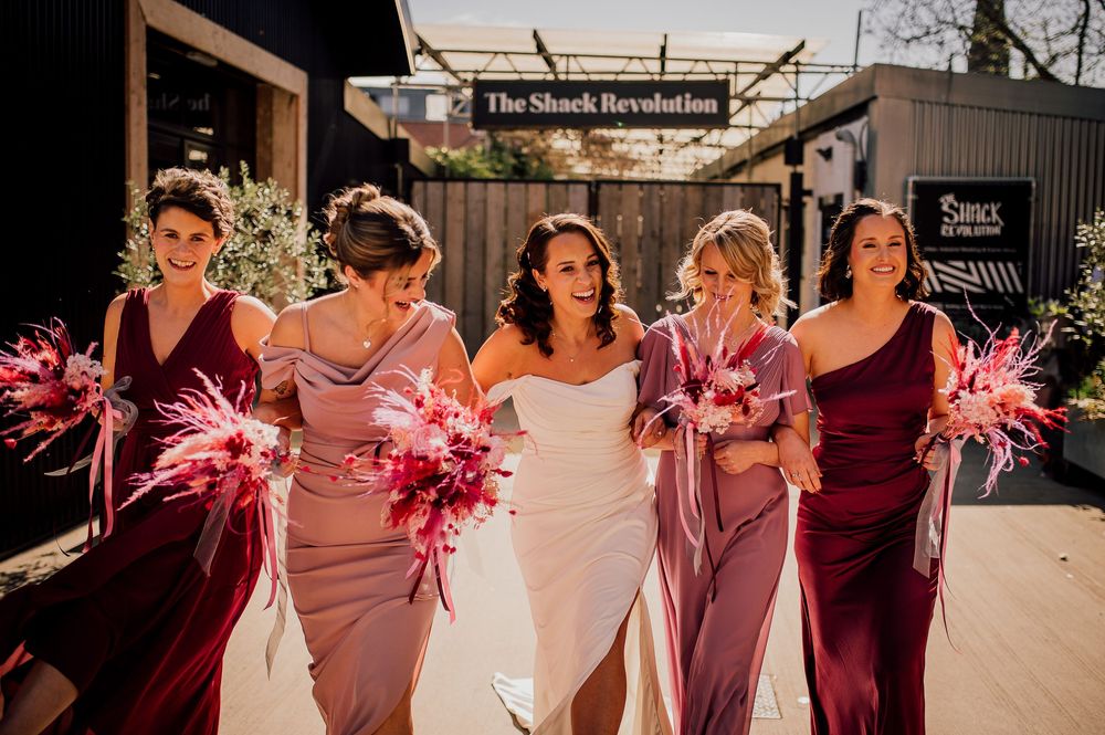 Bridesmaids in pink and burgundy dresses pose together on a sunny day during a wedding celebration.