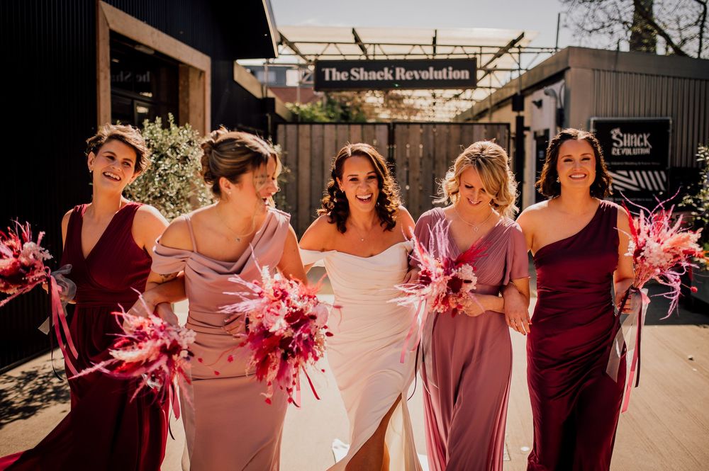 A group in burgundy and pink dresses poses together in outdoor sunlight with feathery pink accessories.