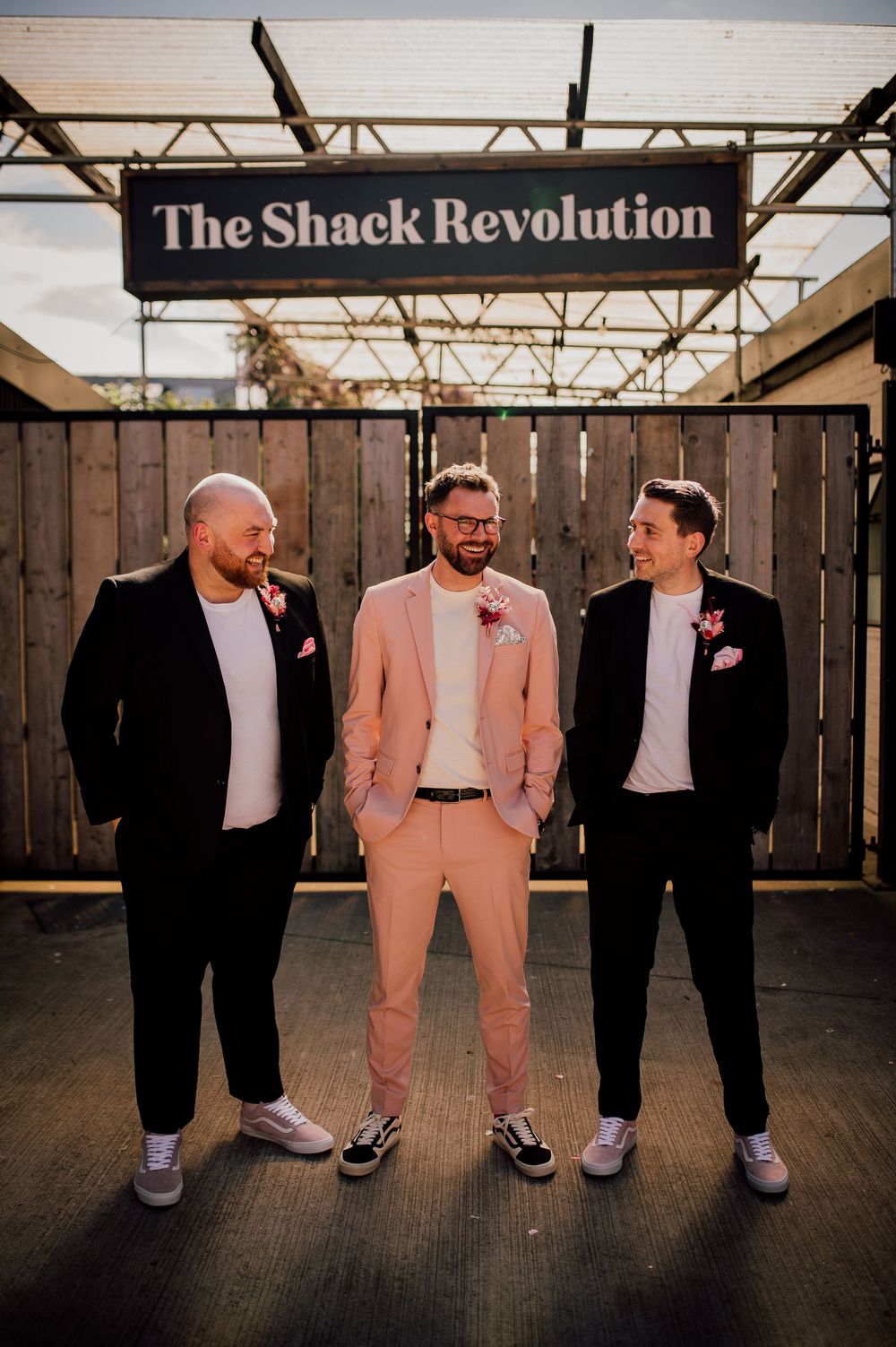 Three men in formal attire stand together against a wooden wall in contrasting black and pink suits.