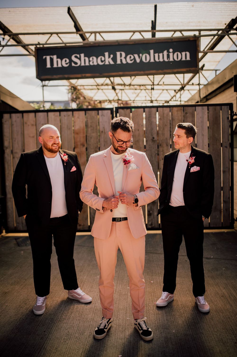 Groomsmen in black suits and one in pink suit pose together showing off their sneakers in a creative wedding photo series.