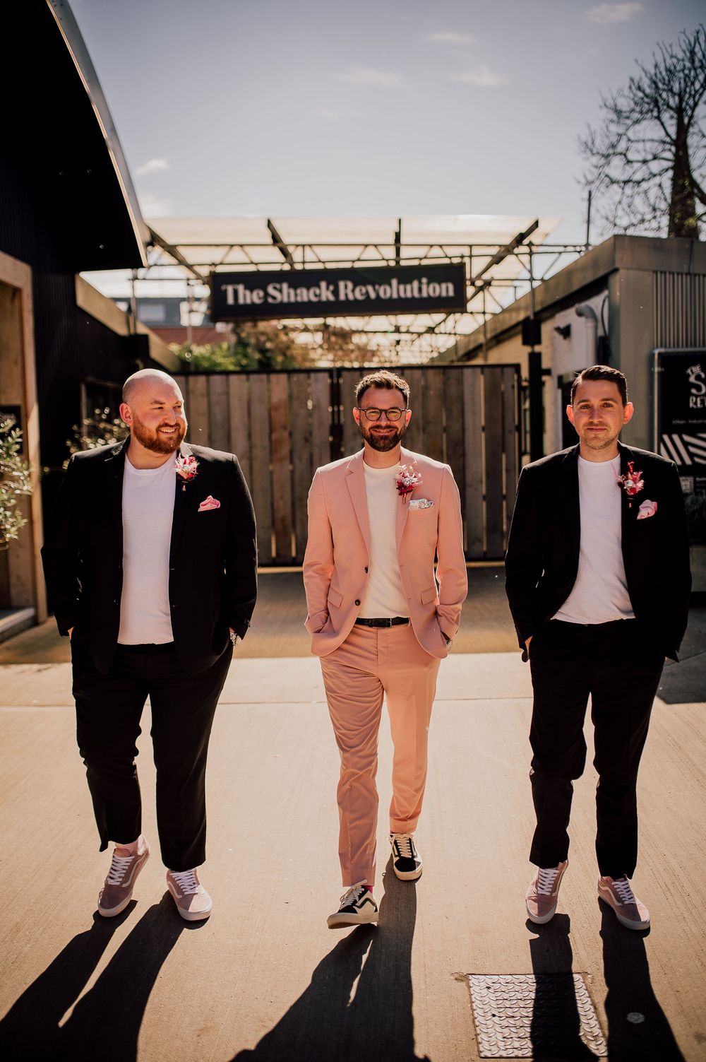 Wedding party members in coordinated black and pink suits pose together in a modern outdoor setting at sunset.