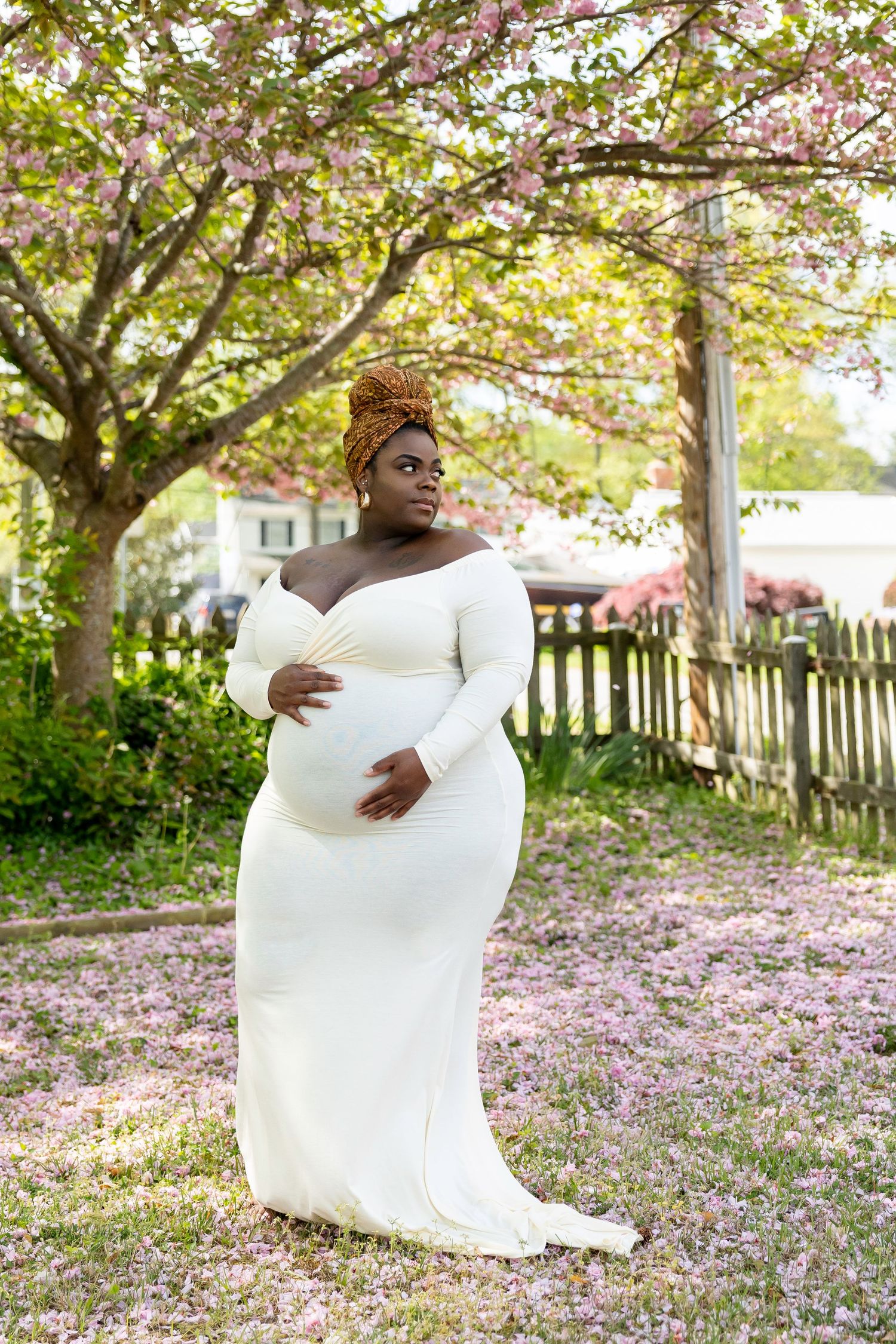 A pregnant Black woman in a white off-shoulder gown poses gracefully under blooming pink cherry blossom trees outdoors.