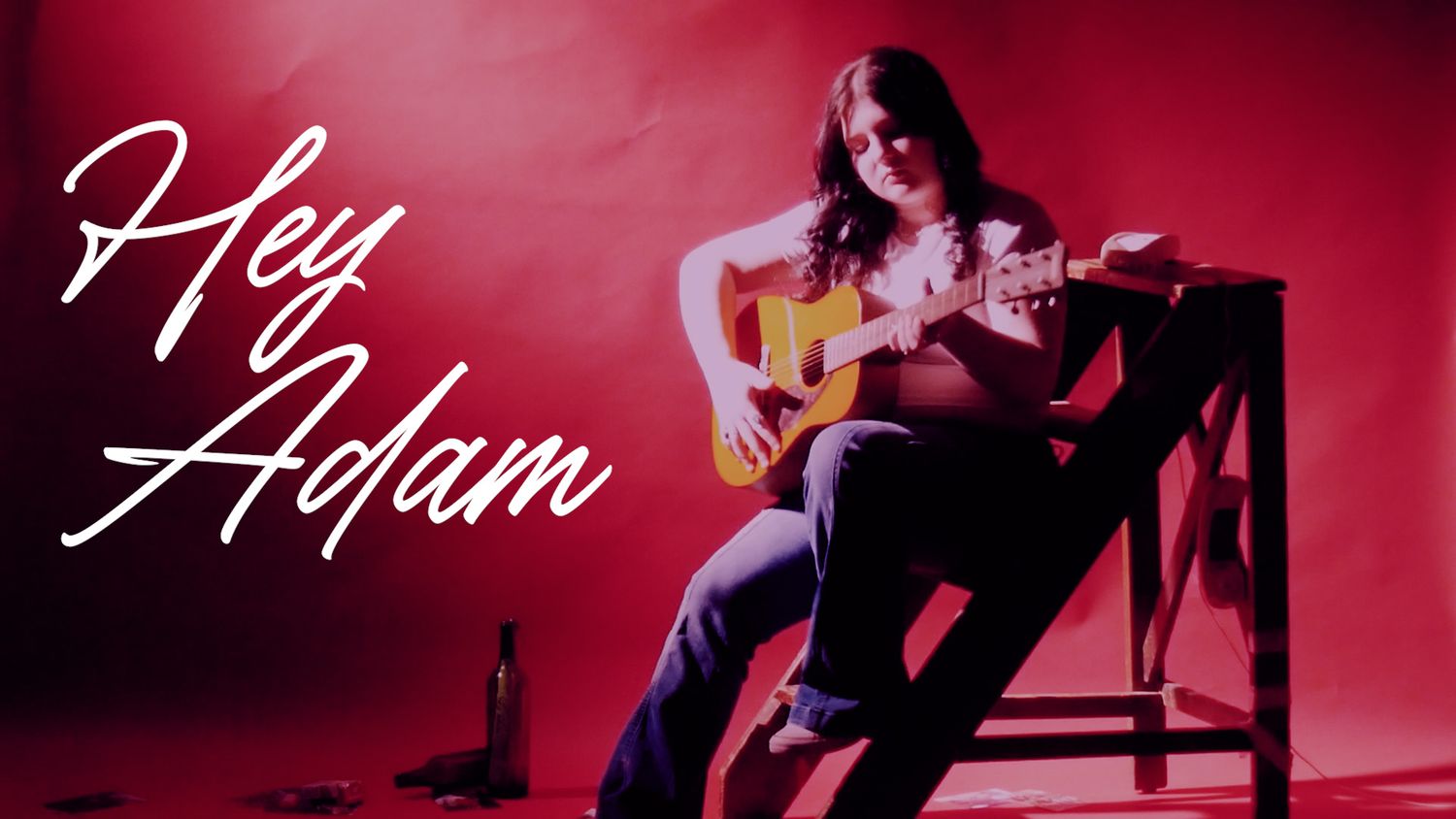 A musician plays an acoustic guitar while sitting on a chair against a red backdrop with 'Hey Adam' text overlay.