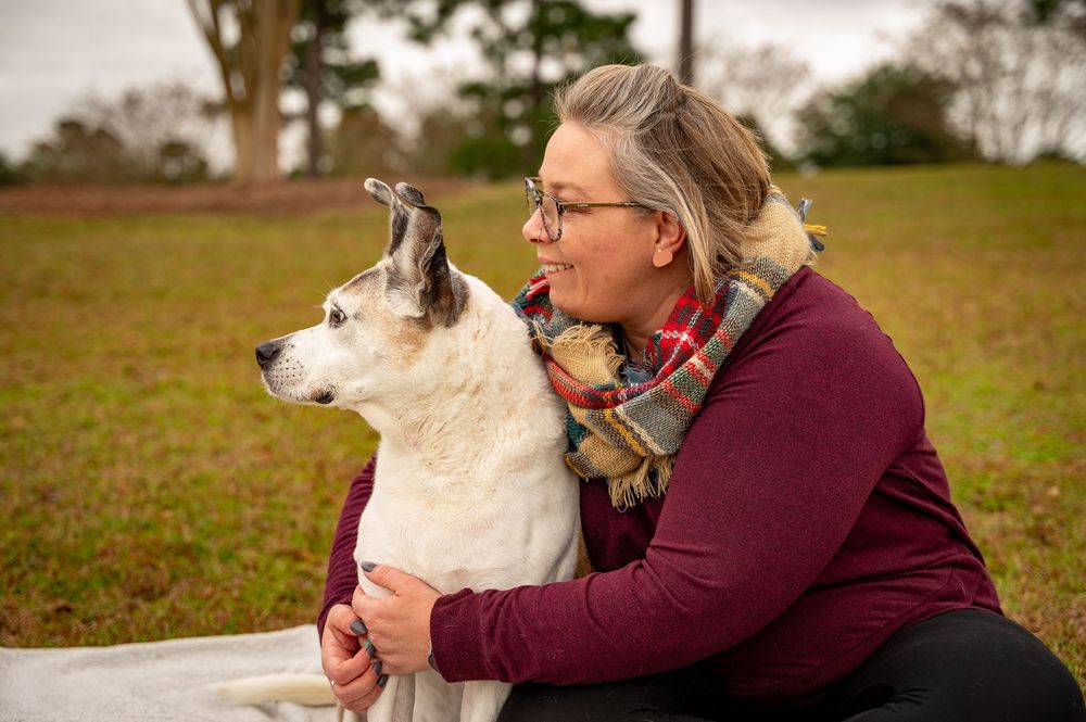 A white dog and person in burgundy sweater share a tender moment outdoors on a grassy field.