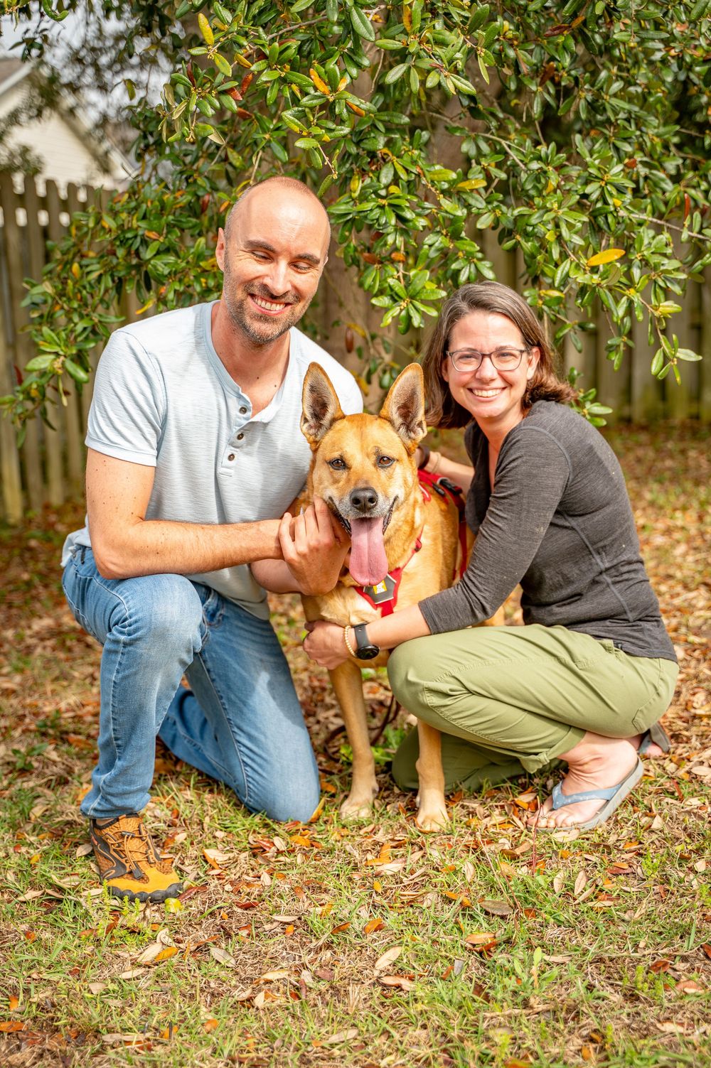 A couple poses with their brown rescue dog on fallen autumn leaves in an outdoor setting.