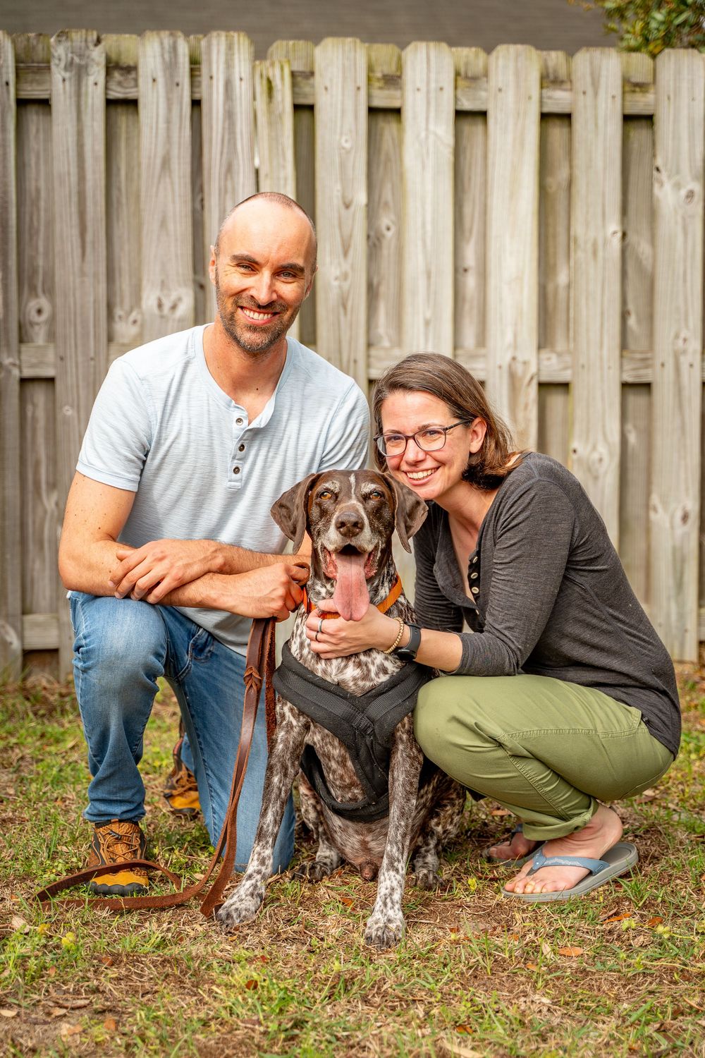 A couple poses with their German Shorthaired Pointer dog in front of a wooden fence in their backyard.