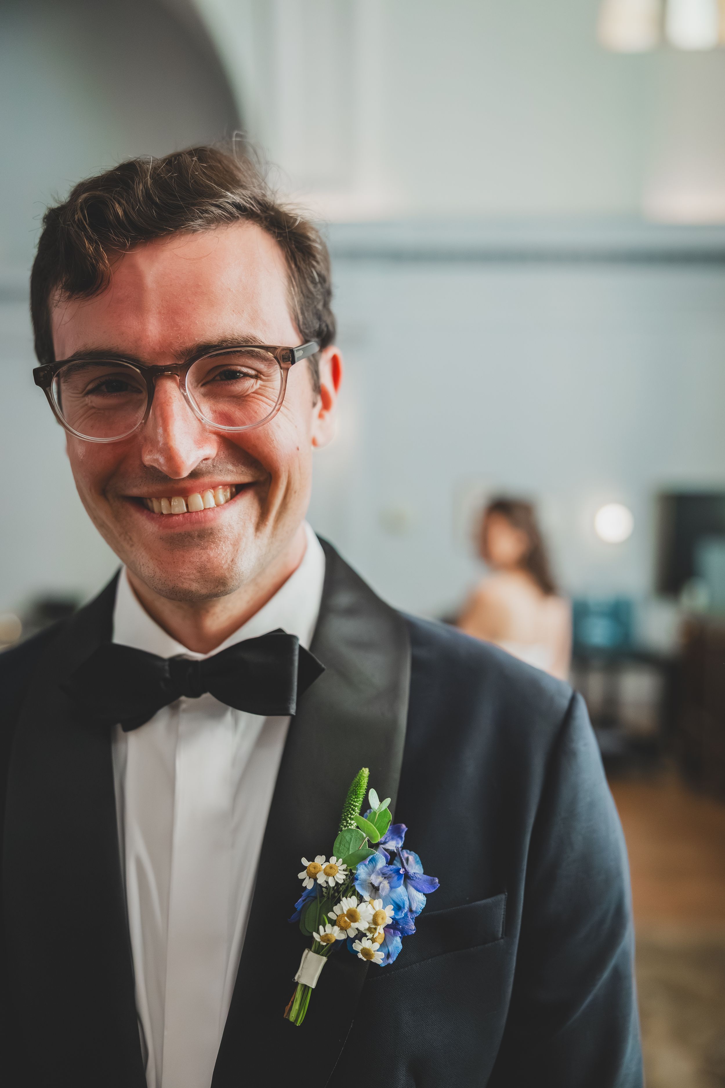 A close-up sequence of a groom wearing glasses and a black tuxedo with a blue and white boutonniere smiling.