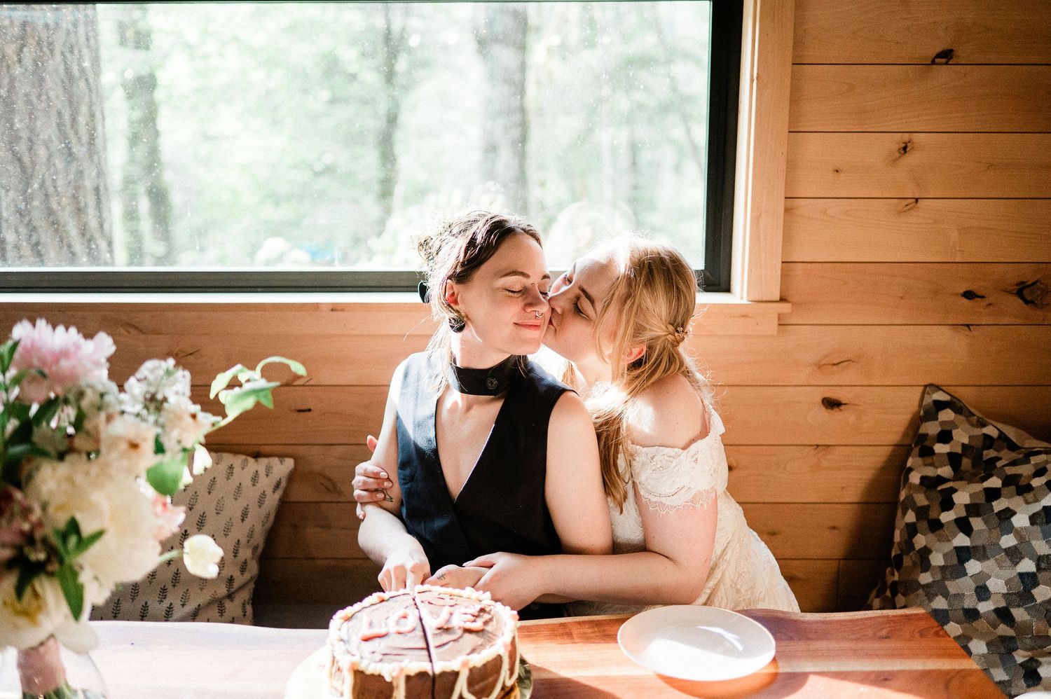 two brides kiss each other on the cheek while cutting cake in a rustic wooden cabin setting.