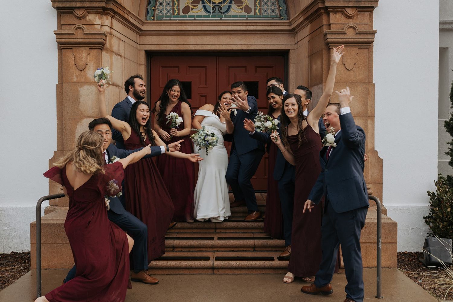 Wedding party celebrates joyfully on church steps with burgundy bridesmaid dresses and formal attire.