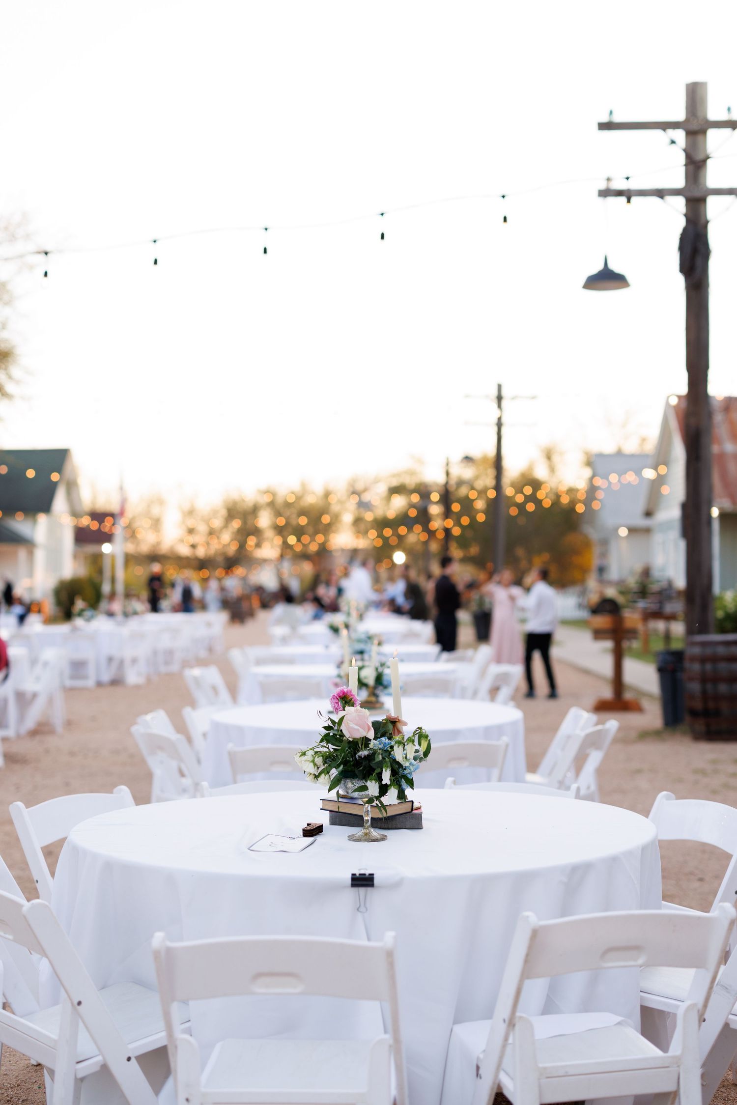 Outdoor wedding reception setup with white round tables, folding chairs and small floral centerpieces at sunset.