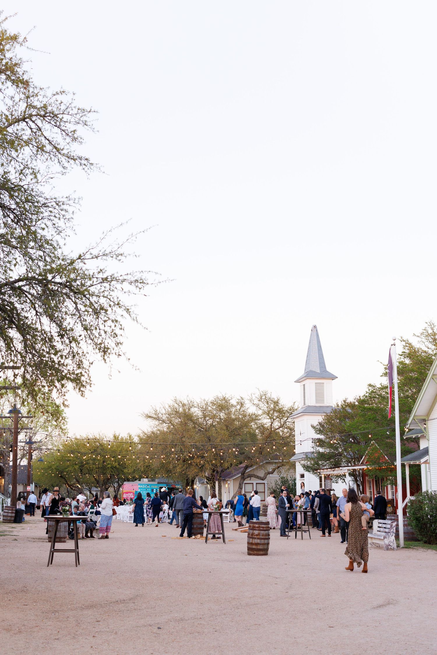Panoramic view of a historic colonial village with white buildings, trees, and visitors walking along dirt paths.