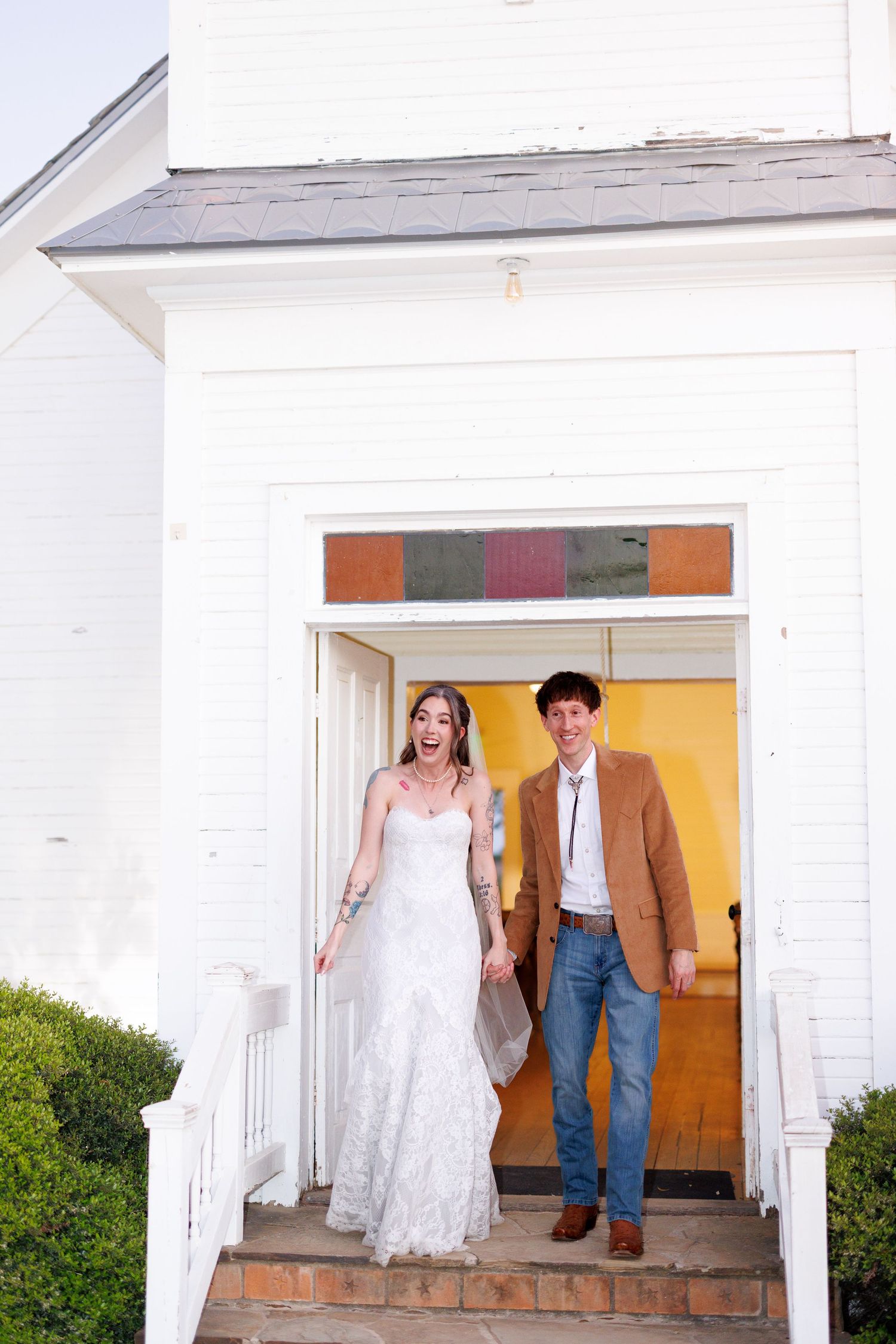 A couple stands in a white doorway entrance, with one person wearing a wedding dress and the other in casual attire.
