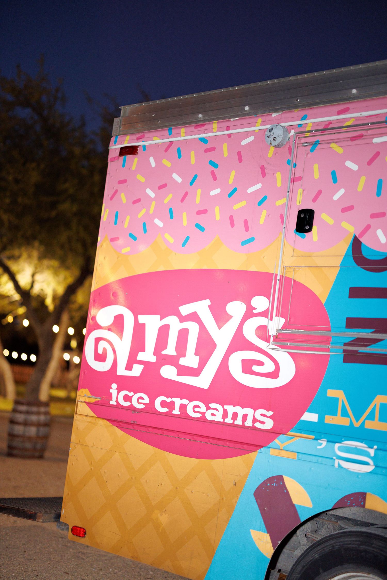 Amy's Ice Cream truck with pink and blue branding displays colorful sprinkle designs against a sunset sky.