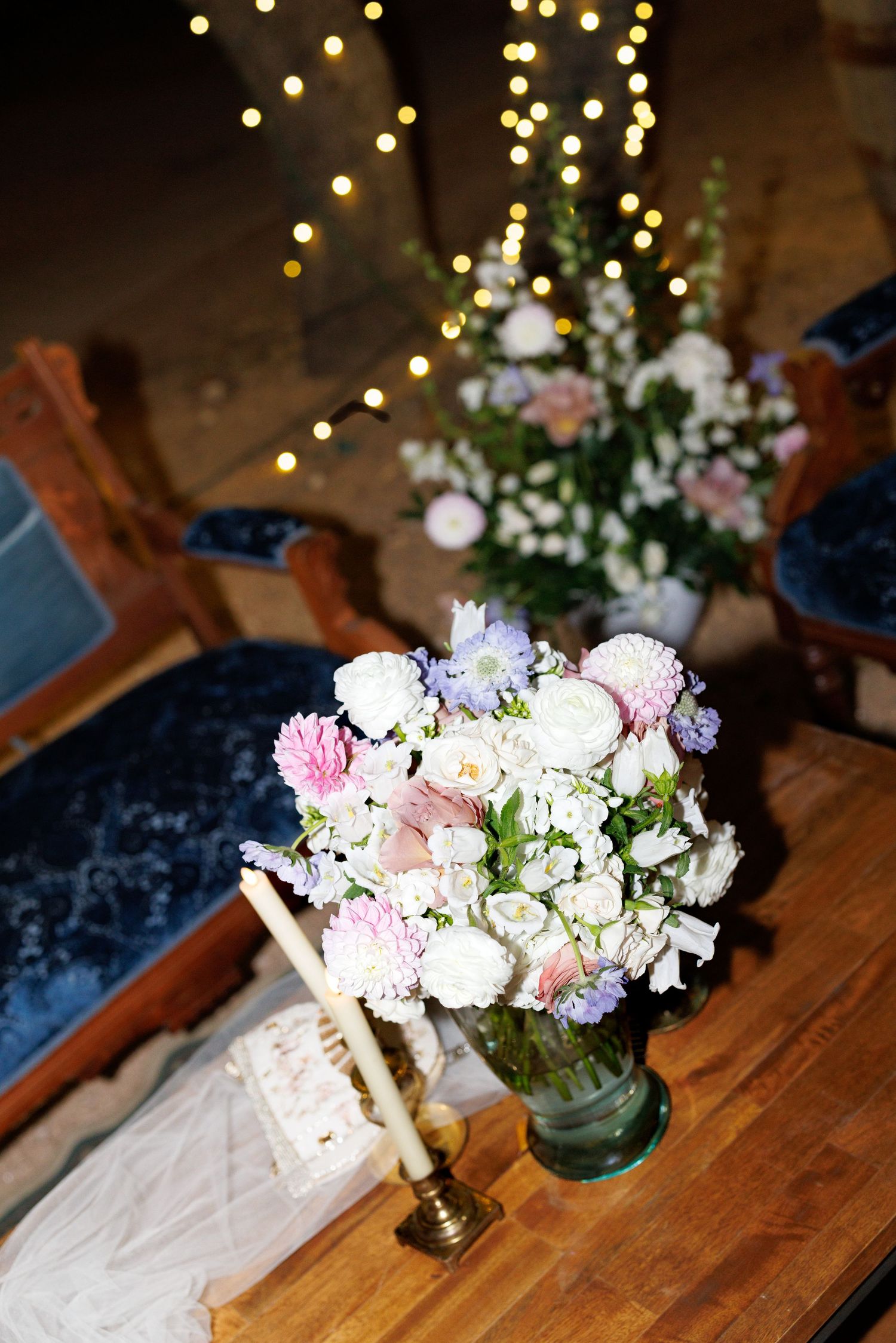 Delicate pink and white flower arrangements on a wooden table with twinkling lights overhead.