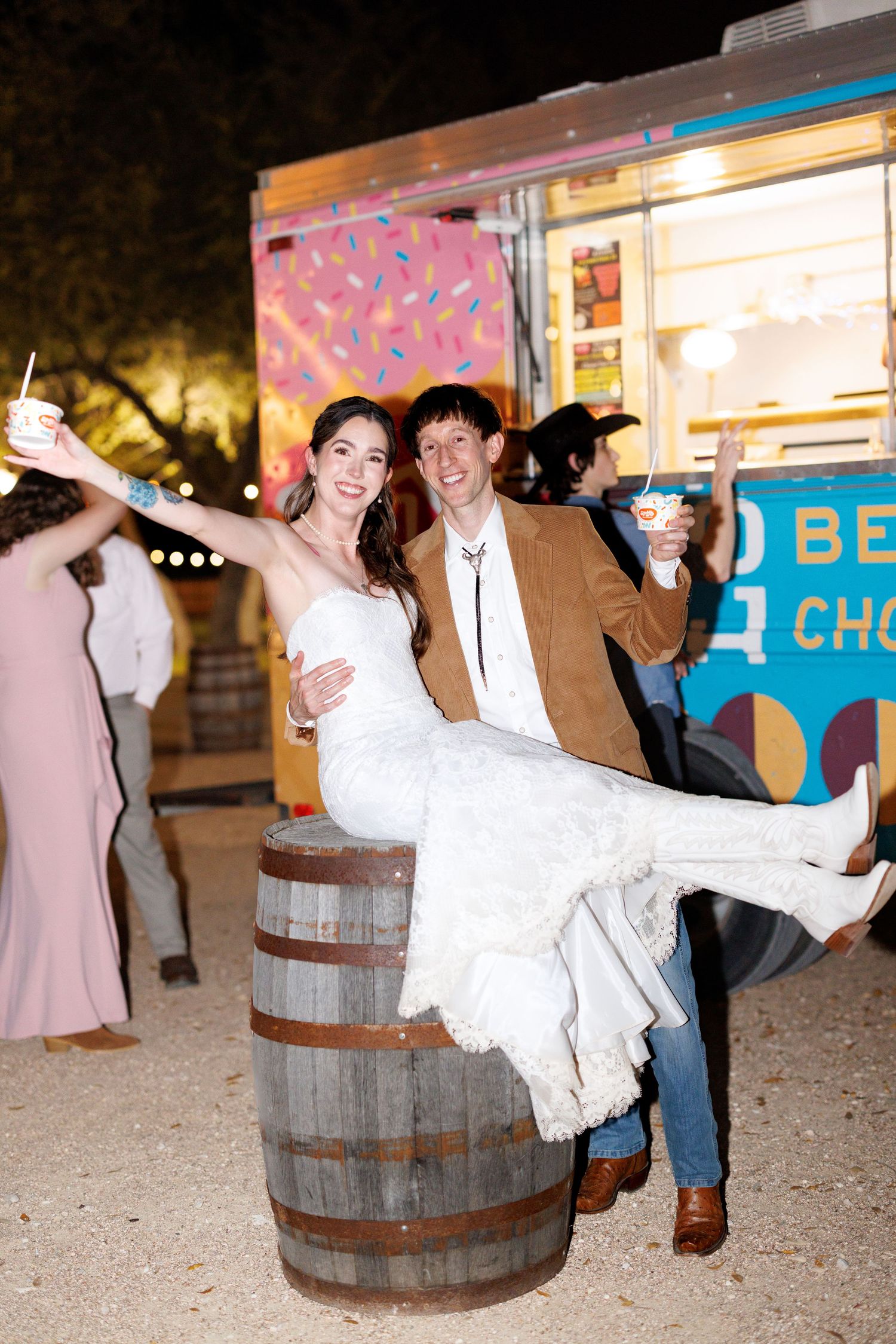 A couple celebrates at a rustic outdoor event, with the bride sitting on a wooden barrel while holding beverages.