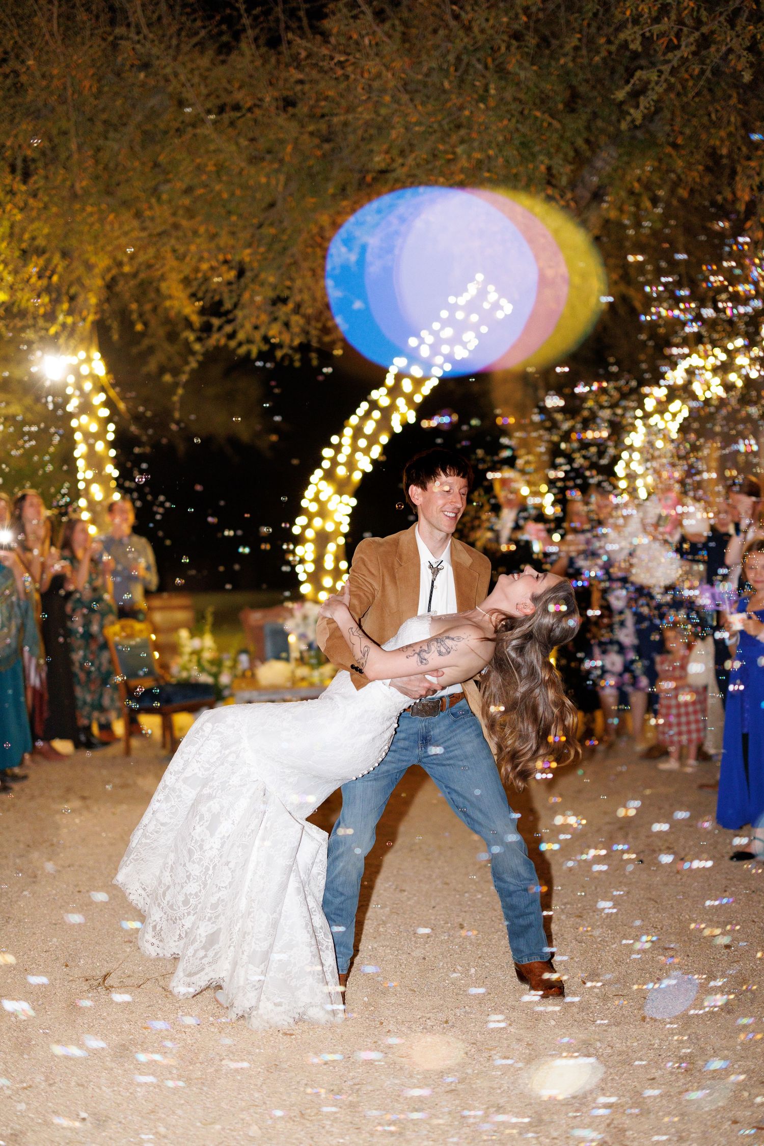 A couple shares a romantic dip during their outdoor wedding celebration with confetti falling around them in the evening.