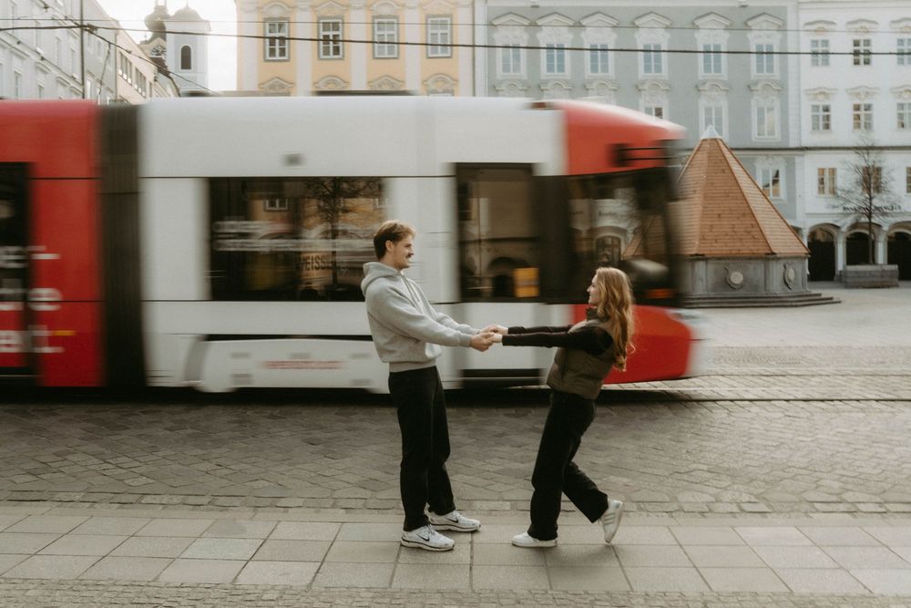 Motion-blurred figures walk past historic European architecture with red trams moving in the background.