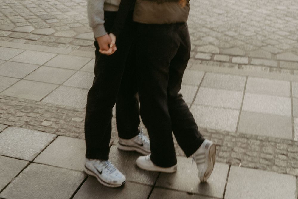 Two pairs of legs in casual pants and sneakers standing close together on a tiled sidewalk.