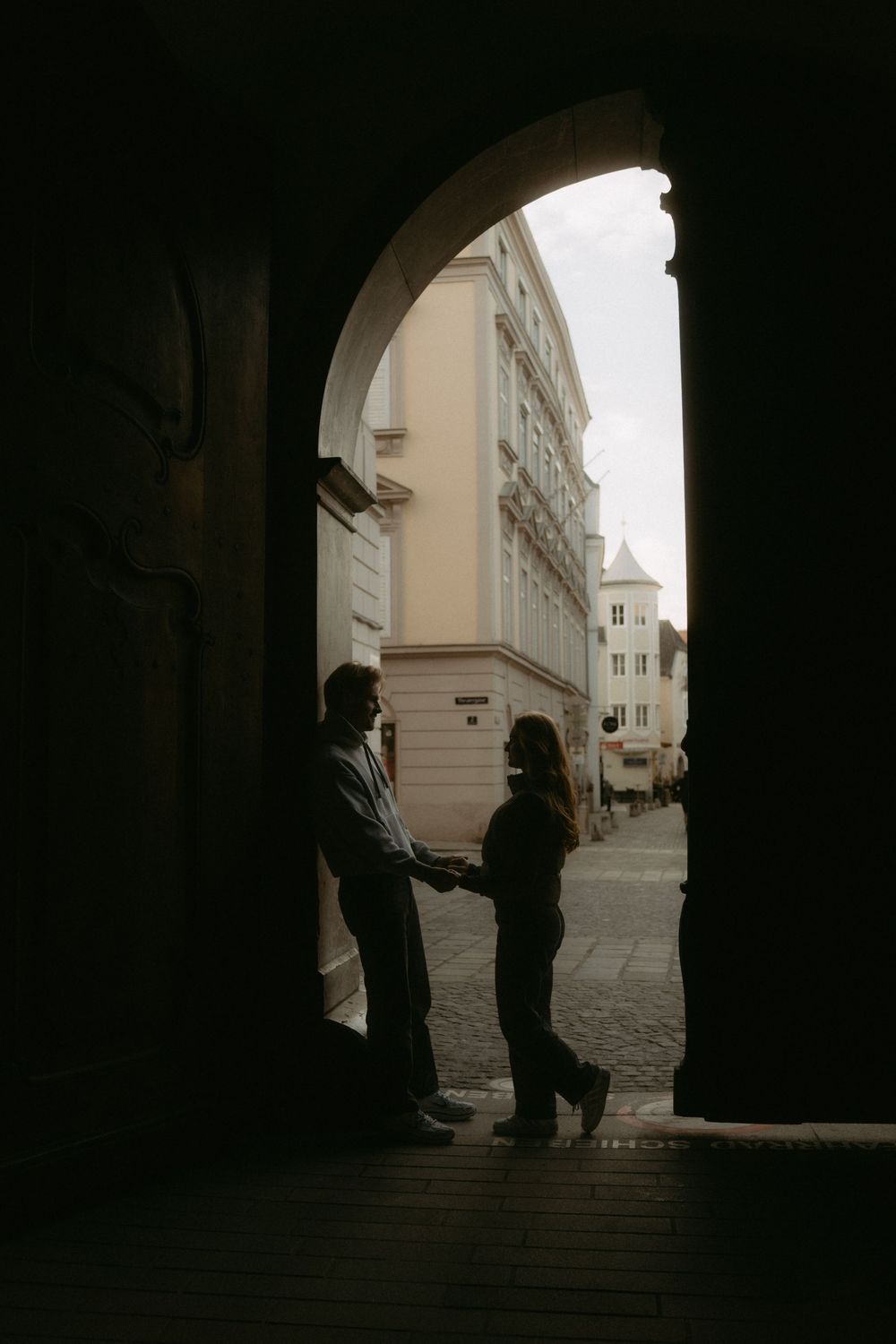 Silhouetted figures stand in a dimly lit archway overlooking a narrow European street.