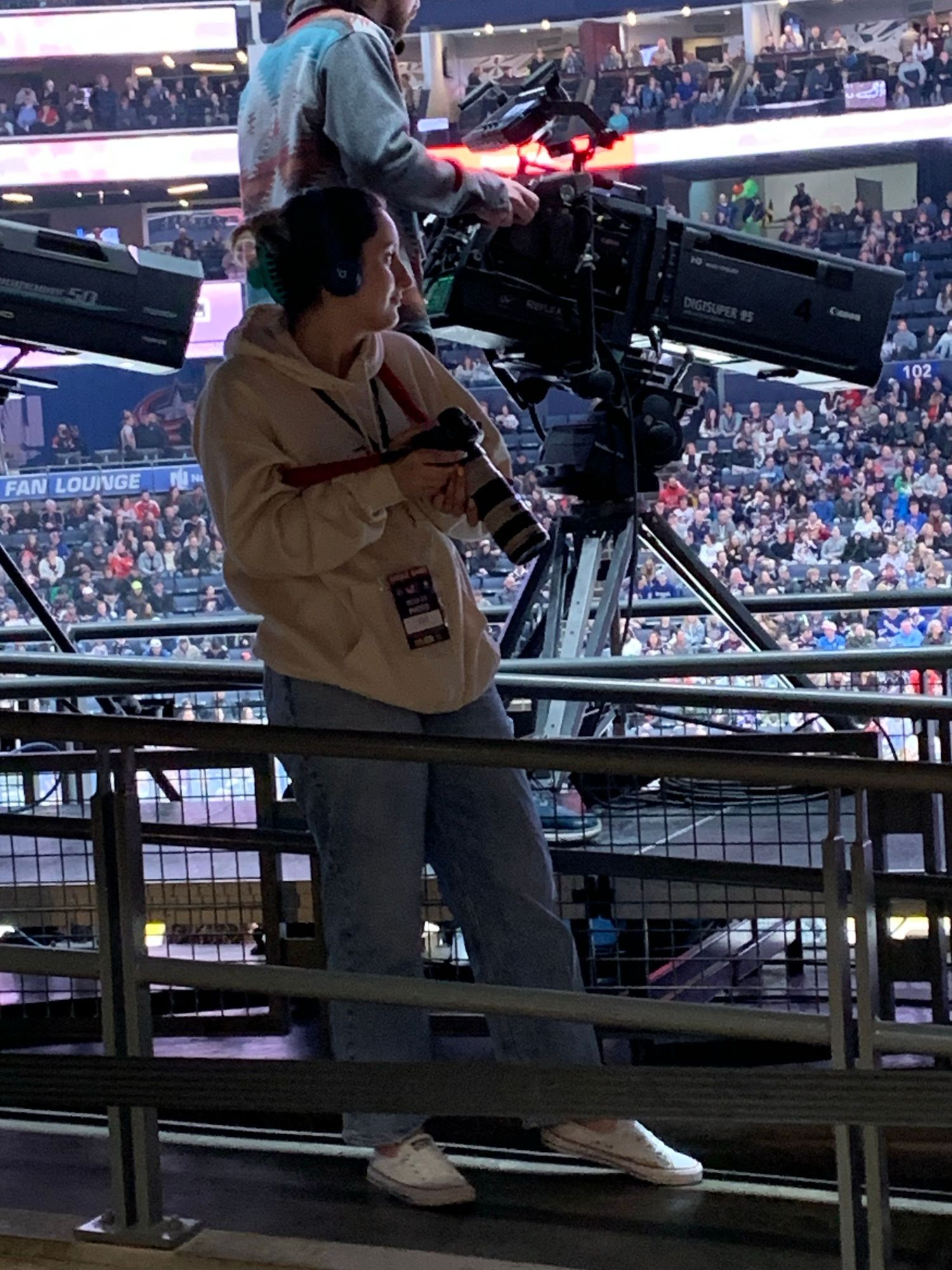 Camera operator with professional broadcast equipment films from elevated position at crowded sports stadium.
