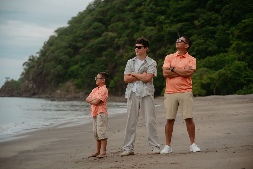 A group of people stand on a scenic beach with lush green cliffs and ocean views in the background.