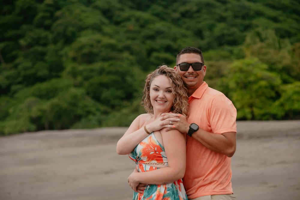 A couple poses together on a beach with lush green mountains in the background while wearing casual summer attire.