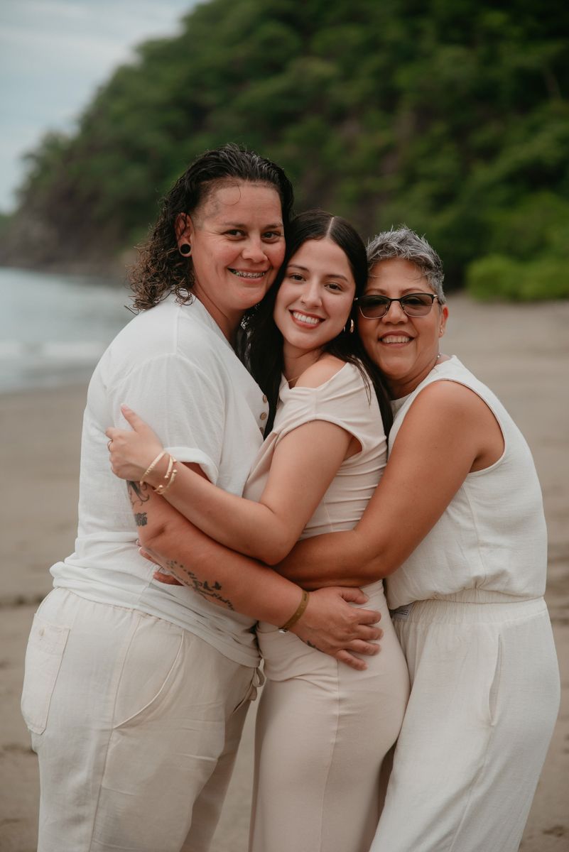 Three people wearing white outfits embrace warmly on a misty beach during a family photoshoot.