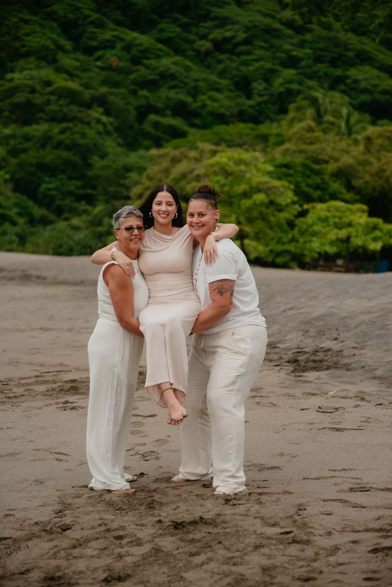 Three people in white outfits share a playful moment together on a sandy beach with lush greenery in background.