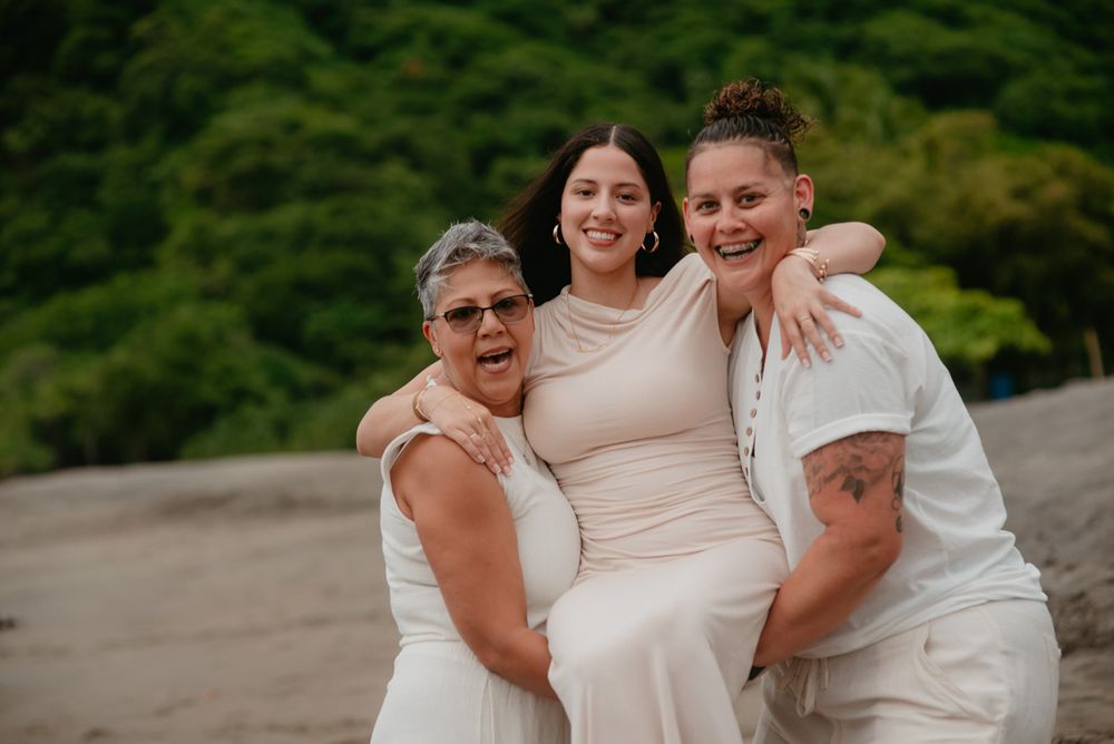 A group in white attire poses together on a sandy beach with lush greenery in the background.