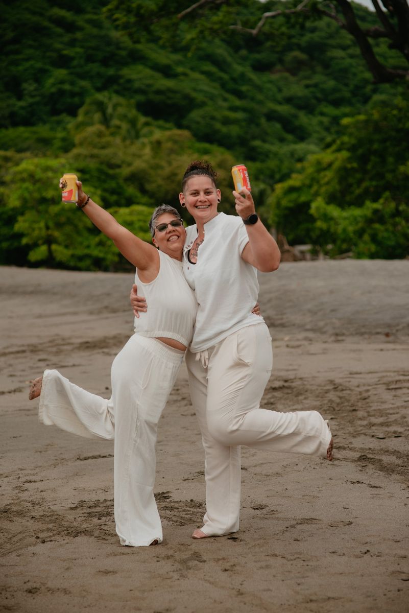 People in white outfits strike fun poses and dance together on a beach with forest backdrop.
