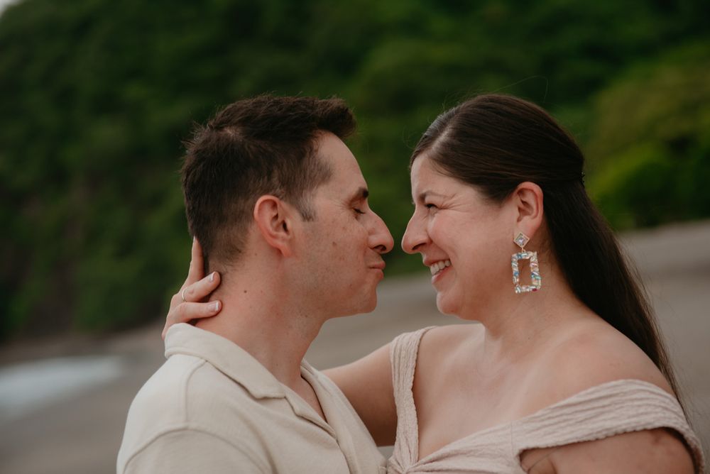 Couple embraces lovingly on sandy beach with tropical foliage and coastline in the distance.