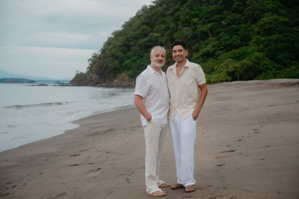 A couple in white outfits walking together on a scenic beach with cliffs and ocean views in the background.