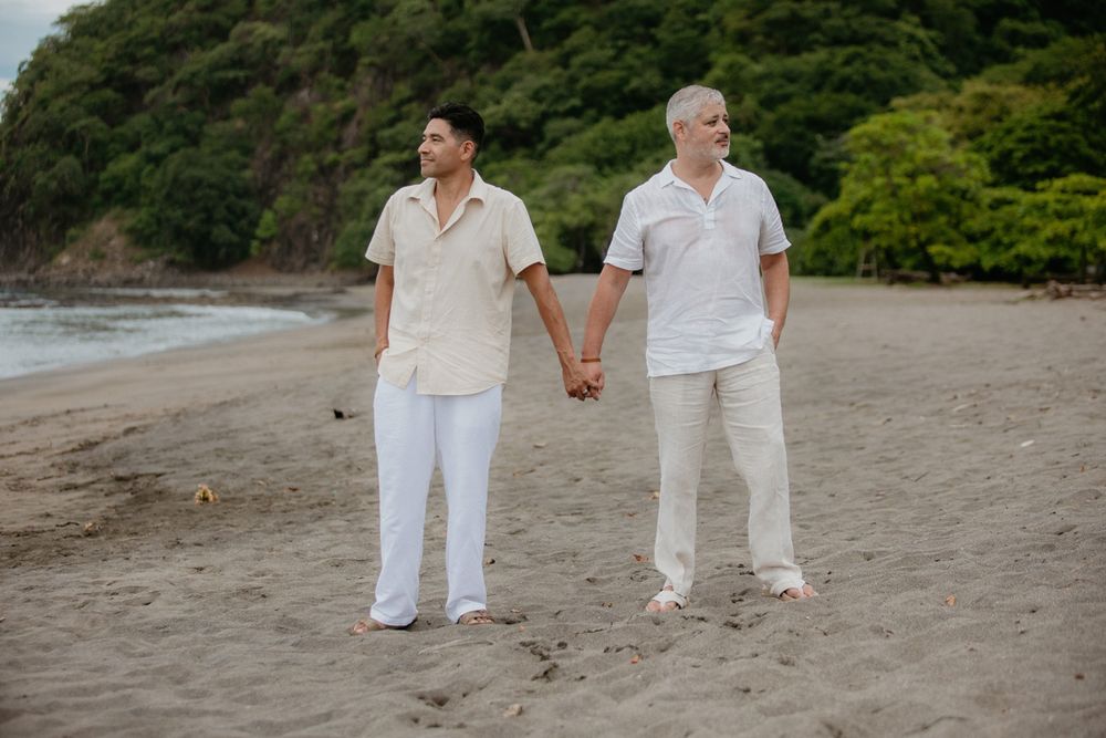 Couple dressed in white walks hand in hand along a peaceful beach with verdant hills behind them.