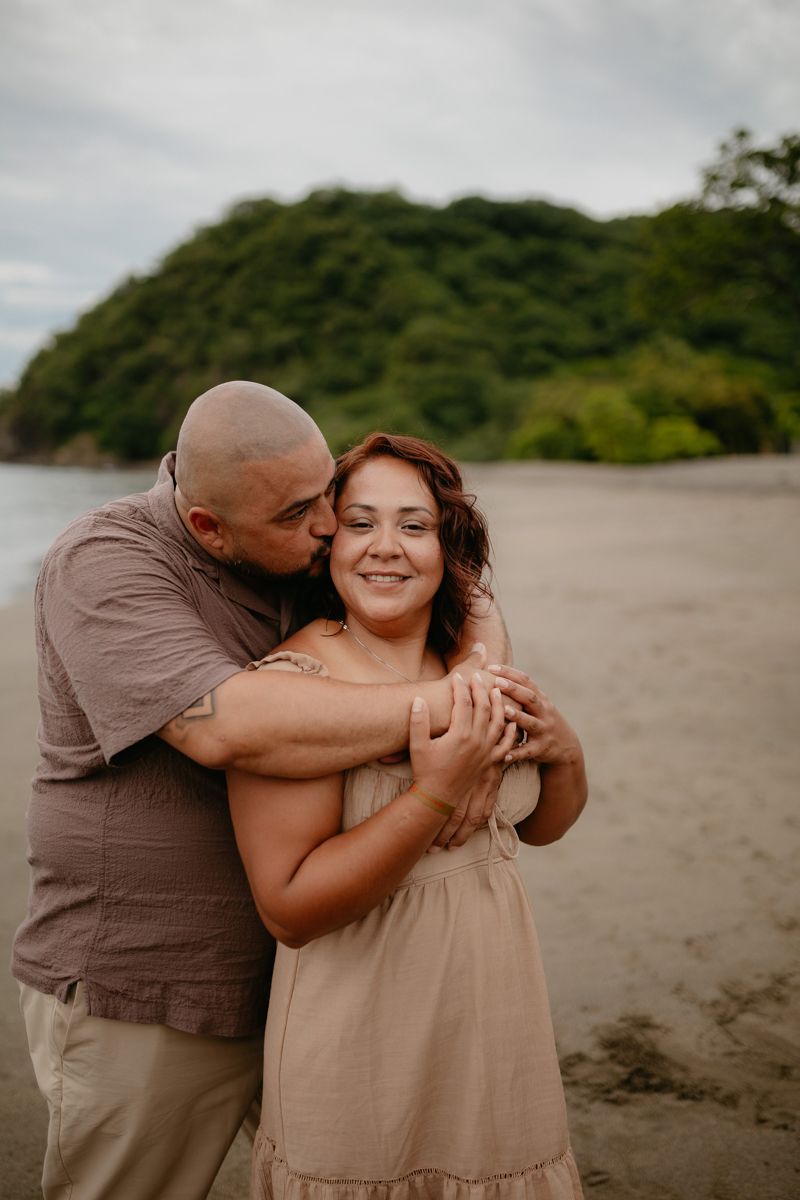 A couple embraces lovingly on a scenic beach with lush greenery and coastline in the background.
